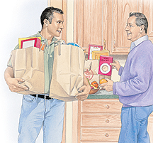 Man bringing grocery bags into kitchen while another man puts groceries away.